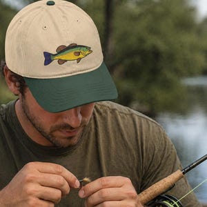 May include: Tan and green baseball cap featuring an embroidered fish design. The cap has a green brim and a matching button on top. The fish is yellow, blue, and brown. A fishing rod is in the foreground.