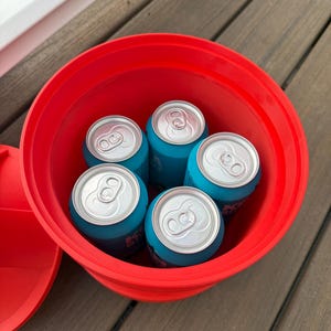 May include: A red plastic bucket filled with five blue beverage cans. The cans have silver tops and are arranged inside the bucket. The bucket is sitting on a wooden surface.