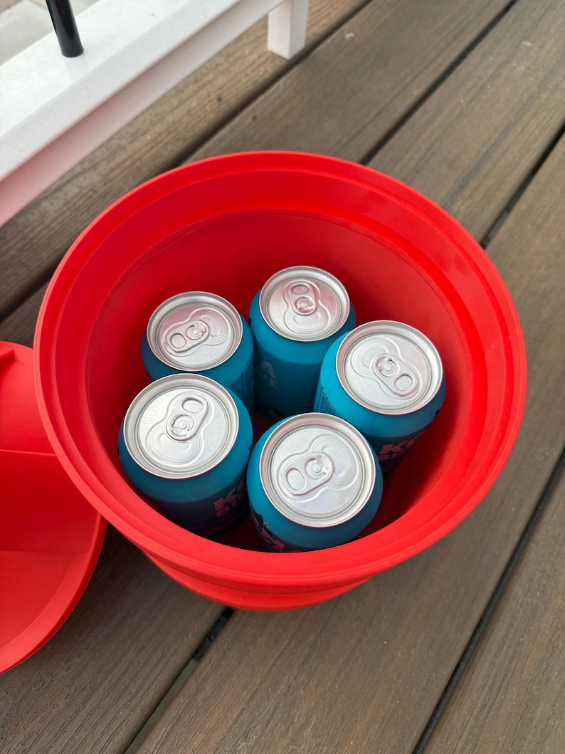 May include: A red plastic bucket filled with five blue beverage cans. The cans have silver tops and are arranged inside the bucket. The bucket is sitting on a wooden surface.