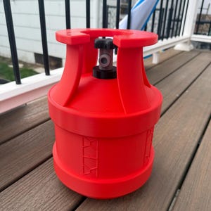 May include: A bright red propane tank with a black valve and a cylindrical body. The tank has a rounded top and bottom with a series of decorative cutouts around the middle. The tank is sitting on a wooden surface.