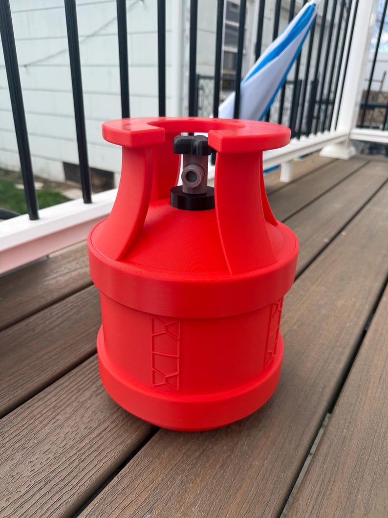 May include: A bright red propane tank with a black valve and a cylindrical body. The tank has a rounded top and bottom with a series of decorative cutouts around the middle. The tank is sitting on a wooden surface.