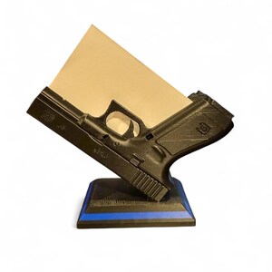 Puede incluir: Un organizador de escritorio negro impreso en 3D con forma de pistola, con un detalle de l&iacute;nea azul en la base. Sostiene un bloc de notas beige en &aacute;ngulo, mostrando los detalles de la pistola.