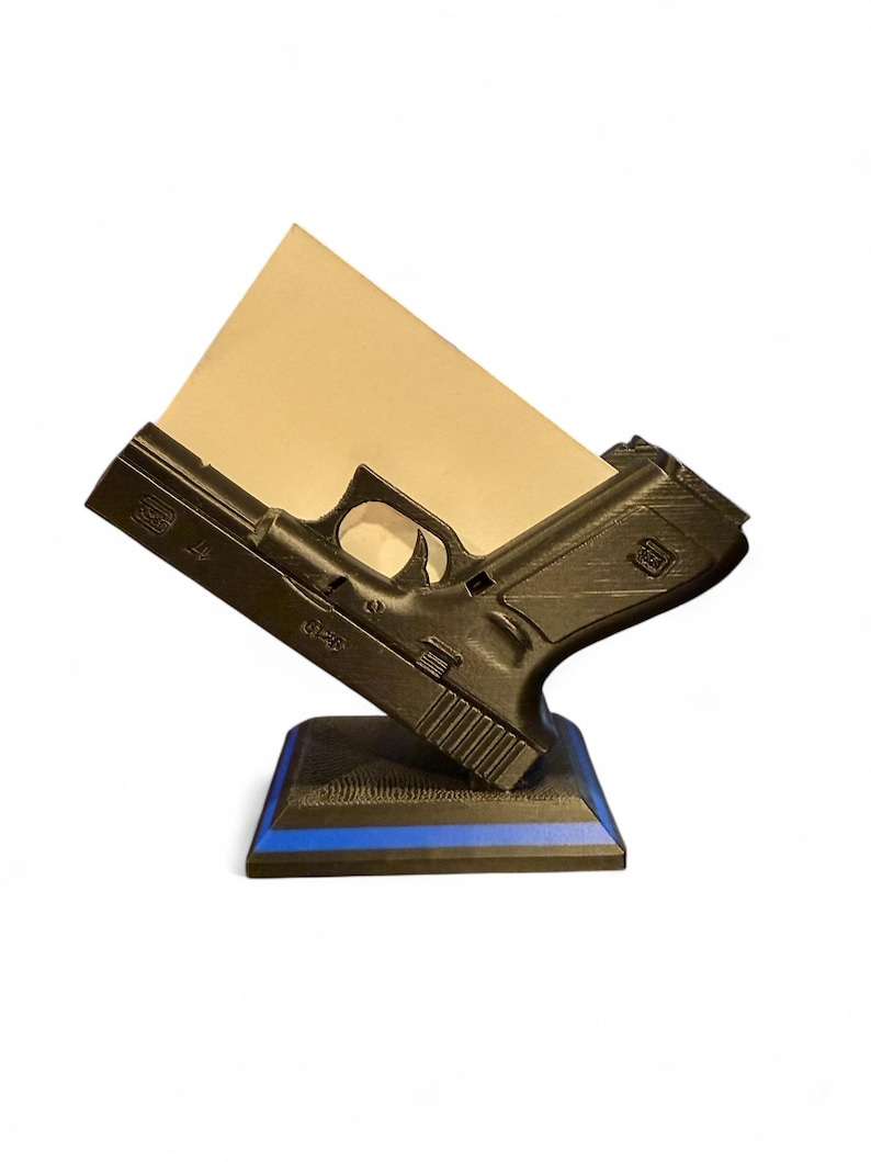 Puede incluir: Un organizador de escritorio negro impreso en 3D con forma de pistola, con un detalle de l&iacute;nea azul en la base. Sostiene un bloc de notas beige en &aacute;ngulo, mostrando los detalles de la pistola.