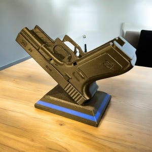 Puede incluir: R&eacute;plica de pistola de color bronce oscuro, montada sobre una base negra con una franja azul. El modelo est&aacute; inclinado hacia arriba, mostrando los detalles del dise&ntilde;o del arma de fuego. La base es rectangular.