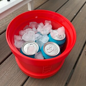 May include: A red, collapsible cooler filled with ice and several blue beverage cans. The cooler is sitting on a wooden deck. The ice cubes are clear and the cans have silver tops.