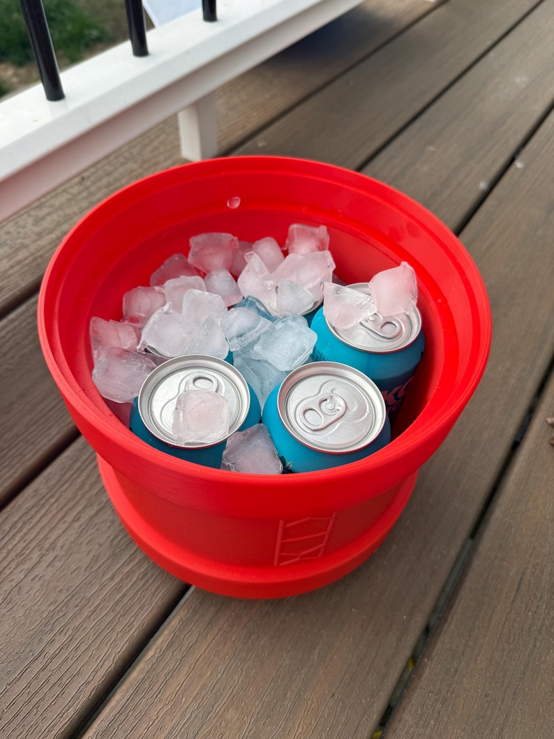 May include: A red, collapsible cooler filled with ice and several blue beverage cans. The cooler is sitting on a wooden deck. The ice cubes are clear and the cans have silver tops.