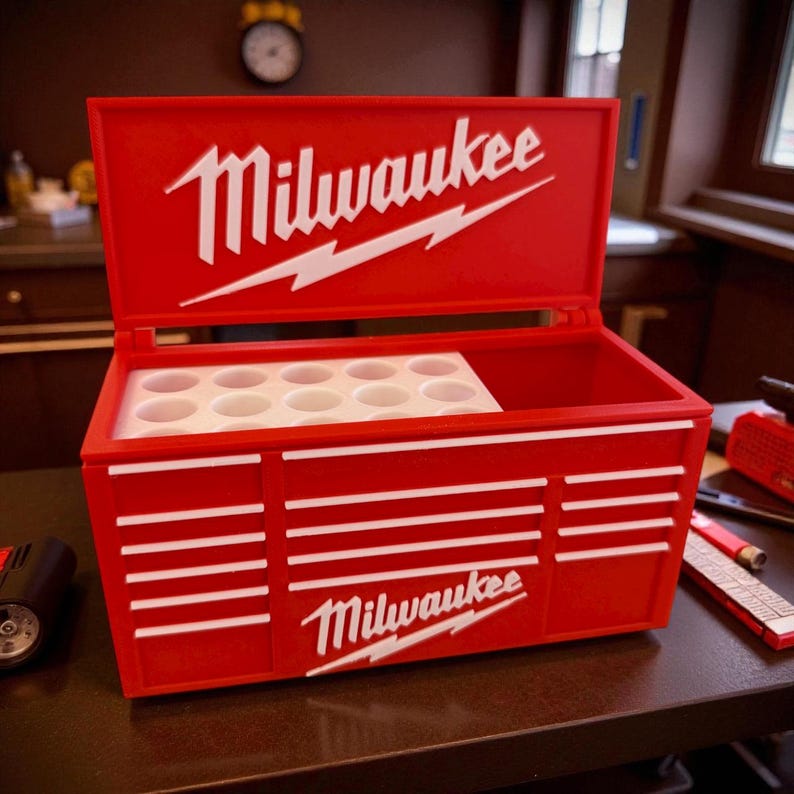 Pu&ograve; includere: Una cassetta degli attrezzi rossa con il logo Milwaukee in bianco. La cassetta ha un coperchio incernierato e strisce orizzontali bianche sul davanti. All'interno, c'&egrave; un inserto bianco con pi&ugrave; aperture circolari.