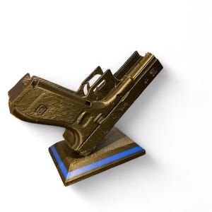 Puede incluir: Un modelo de pistola de color dorado apoyado sobre un soporte marr&oacute;n con una franja azul. El modelo presenta ranuras y texturas detalladas, lo que sugiere un dise&ntilde;o impreso en 3D. El soporte est&aacute; inclinado, proporcionando una presentaci&oacute;n din&aacute;mica.
