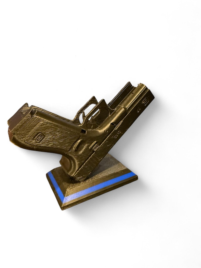 Puede incluir: Un modelo de pistola de color dorado apoyado sobre un soporte marr&oacute;n con una franja azul. El modelo presenta ranuras y texturas detalladas, lo que sugiere un dise&ntilde;o impreso en 3D. El soporte est&aacute; inclinado, proporcionando una presentaci&oacute;n din&aacute;mica.