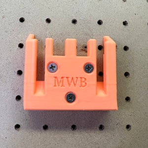 Puede incluir: Soporte de pared de pl&aacute;stico naranja con cuatro ranuras y el texto "MWB" impreso en la parte delantera.