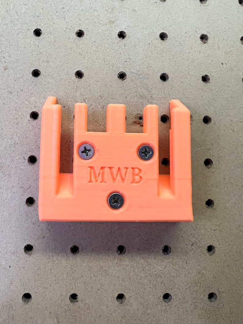 Puede incluir: Soporte de pared de pl&aacute;stico naranja con cuatro ranuras y el texto "MWB" impreso en la parte delantera.