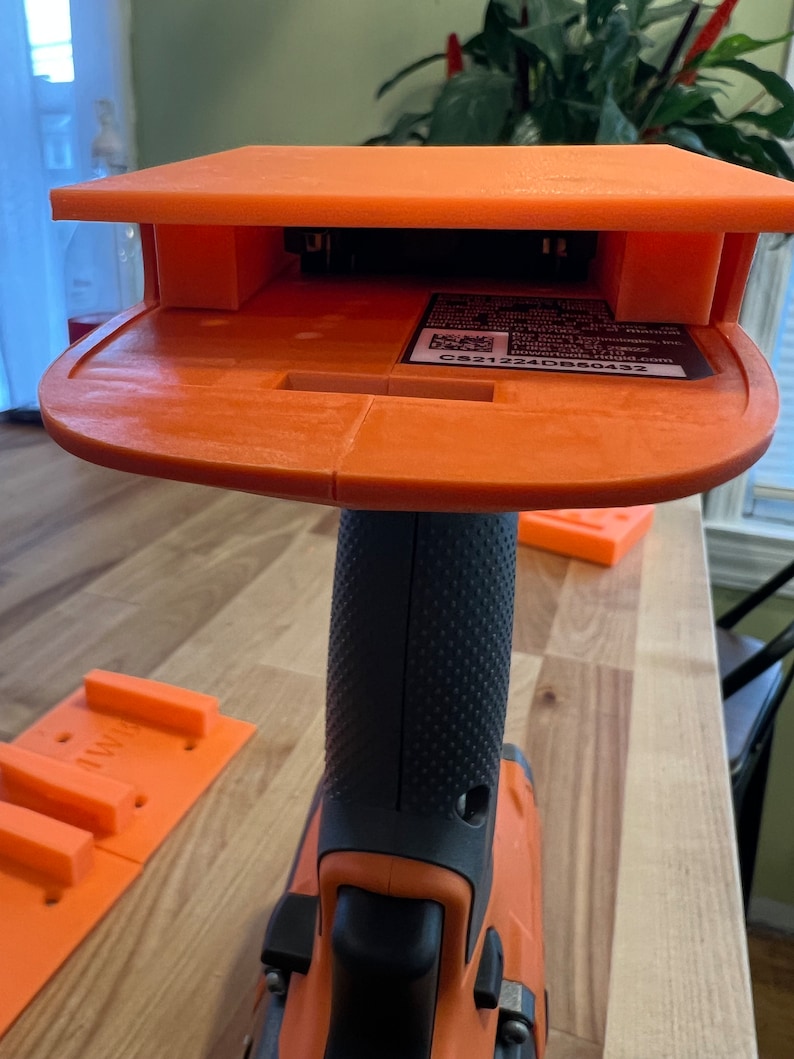 Puede incluir: Soporte de taladro impreso en 3D de color naranja con un taladro negro conectado. El soporte tiene un compartimento para guardar una bater&iacute;a. La bater&iacute;a es visible dentro del compartimento. El soporte tiene una etiqueta con el texto "CS21224DB50432".
