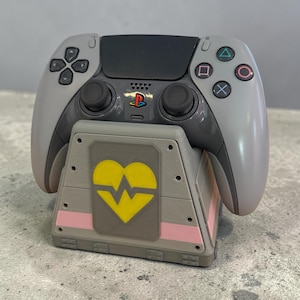 以下が含まれることがあります： グレーと黒のPlayStation 5コントローラーが、カスタムデザインのスタンドに置かれています。スタンドはグレーで、ピンクと黄色のアクセントがあり、ハート型のデザインが施されています。コントローラーにはPlayStationのロゴがあります。