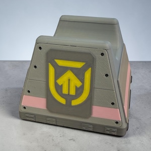 以下が含まれることがあります： 前面に黄色の矢印のシンボルが付いた、灰色の幾何学的な箱。箱にはピンクと茶色のアクセントがあります。デザインは3Dプリントのようで、レイヤーラインが見えます。箱はユニークで角張った形をしています。