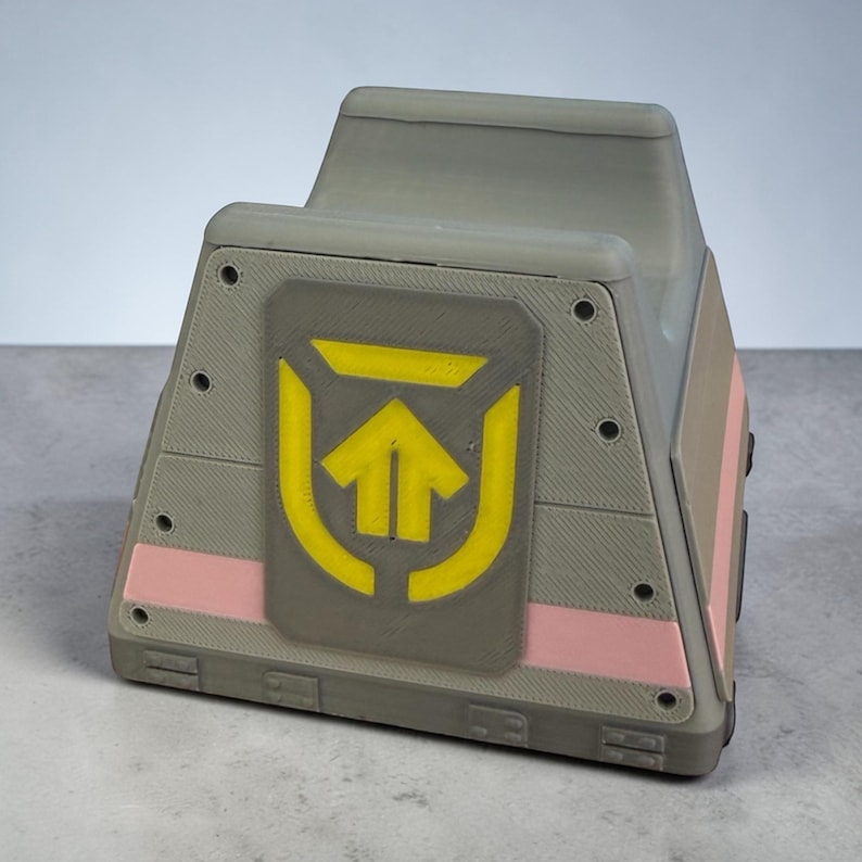 以下が含まれることがあります： 前面に黄色の矢印のシンボルが付いた、灰色の幾何学的な箱。箱にはピンクと茶色のアクセントがあります。デザインは3Dプリントのようで、レイヤーラインが見えます。箱はユニークで角張った形をしています。