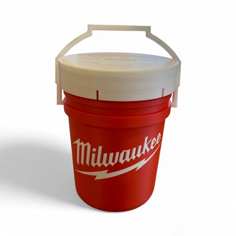 May include: A red bucket with a white lid and handle. The bucket has the word "Milwaukee" in white script with a lightning bolt design. The handle is arched and attached to the sides of the bucket.