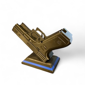 Puede incluir: R&eacute;plica de pistola de color dorado, montada sobre una base negra con una franja azul. La pistola modelo est&aacute; inclinada hacia arriba, mostrando sus detalles. El fondo es blanco liso.
