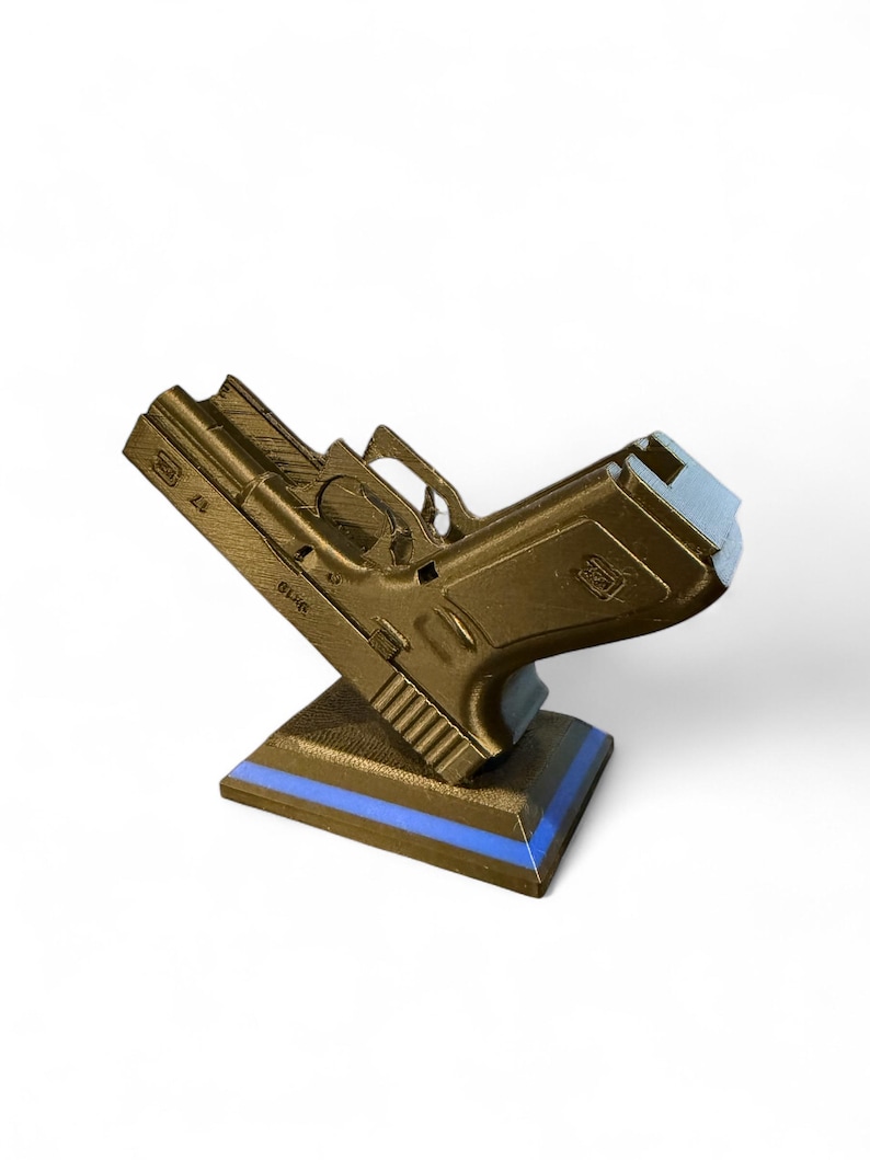 Puede incluir: R&eacute;plica de pistola de color dorado, montada sobre una base negra con una franja azul. La pistola modelo est&aacute; inclinada hacia arriba, mostrando sus detalles. El fondo es blanco liso.