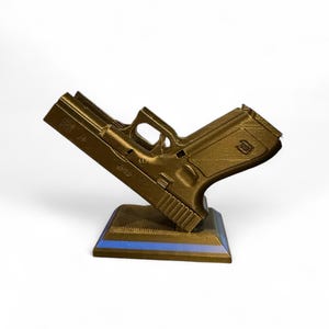 Puede incluir: R&eacute;plica dorada impresa en 3D de una pistola, apoyada sobre una base oscura con una franja azul. La pistola est&aacute; en &aacute;ngulo, mostrando sus detalles sobre un fondo blanco. El modelo tiene marcas visibles.