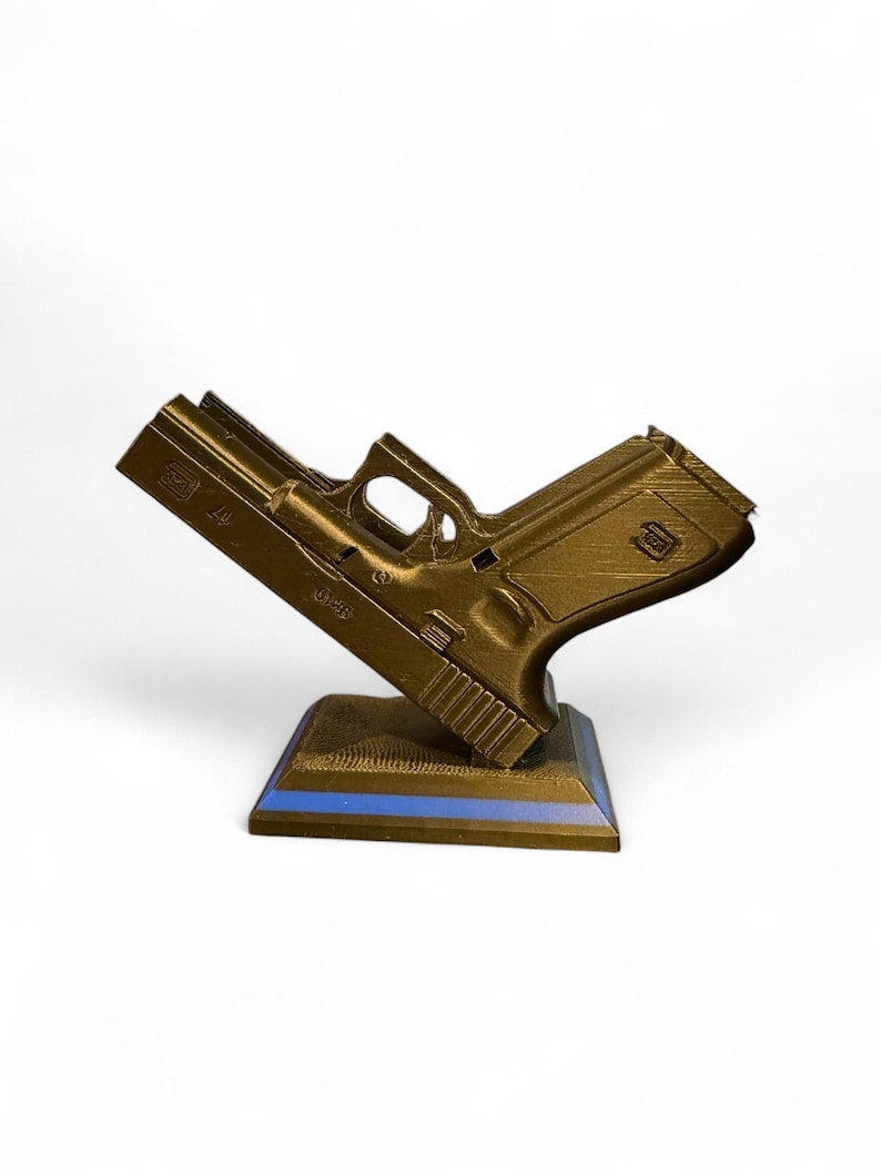Puede incluir: R&eacute;plica dorada impresa en 3D de una pistola, apoyada sobre una base oscura con una franja azul. La pistola est&aacute; en &aacute;ngulo, mostrando sus detalles sobre un fondo blanco. El modelo tiene marcas visibles.