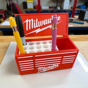Pu&ograve; includere: Un portapenne rosso a forma di cassetta degli attrezzi con il logo "Milwaukee" in bianco. Contiene una penna gialla, un pennarello nero e una penna rosa. La cassetta degli attrezzi ha un interno bianco con pi&ugrave; fori per penne.