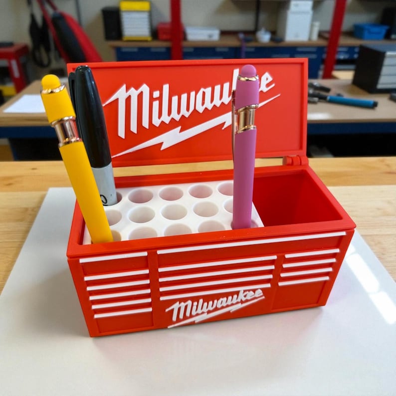 Pu&ograve; includere: Un portapenne rosso a forma di cassetta degli attrezzi con il logo "Milwaukee" in bianco. Contiene una penna gialla, un pennarello nero e una penna rosa. La cassetta degli attrezzi ha un interno bianco con pi&ugrave; fori per penne.