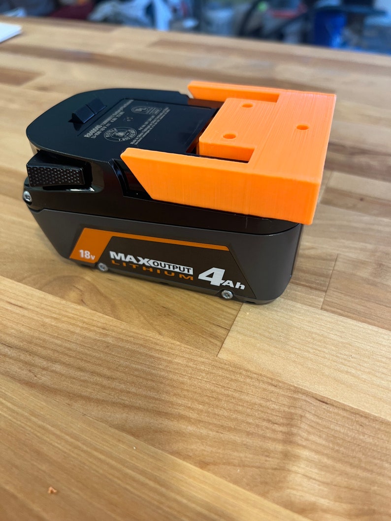 Puede incluir: Un soporte de bater&iacute;a impreso en 3D de color naranja para una bater&iacute;a de iones de litio negra de 18 V con una capacidad de 4 Ah. La bater&iacute;a tiene el texto "MAX OUTPUT LITHIUM" impreso en ella.