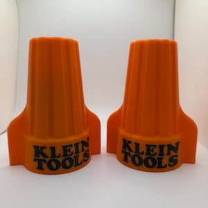 Könnte beinhalten: Zwei orangefarbene Klein Tools-Drahtverbinder. Jeder Verbinder hat eine gerippte Oberseite und den Markennamen in schwarzer Schrift. Die Verbinder haben eine ausgestellte Basis mit einem flügelartigen Design.