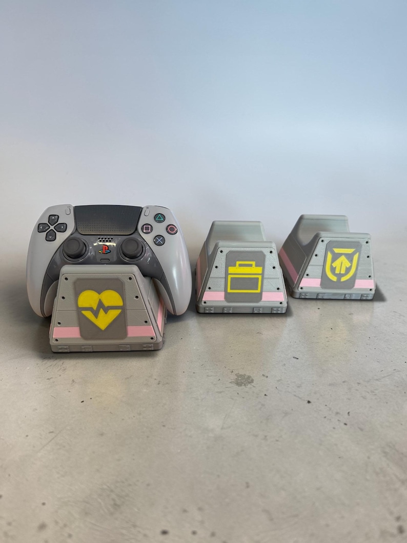 以下が含まれることがあります： 白とグレーのゲームコントローラーが、黄色のハートのグラフィックが付いたグレーのベースの上に置かれています。ピンクのアクセントと、ブリーフケースと矢印の黄色のグラフィックが付いた、2つの追加のグレーのベースも表示されます。ゲームアイテム。