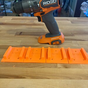 Puede incluir: Un taladro inal&aacute;mbrico negro y naranja con el nombre de la marca "RIDGID" impreso en el lateral. El taladro est&aacute; sobre una superficie de madera con cuatro soportes de pared de pl&aacute;stico naranja etiquetados como "MWB".
