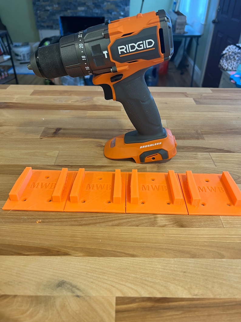 Puede incluir: Un taladro inal&aacute;mbrico negro y naranja con el nombre de la marca "RIDGID" impreso en el lateral. El taladro est&aacute; sobre una superficie de madera con cuatro soportes de pared de pl&aacute;stico naranja etiquetados como "MWB".