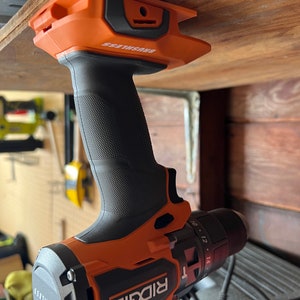 Puede incluir: Un taladro inal&aacute;mbrico naranja y negro con el nombre de la marca "RIDGID" y el texto "BRUSHLESS" en el lateral. El taladro est&aacute; colgado de una estanter&iacute;a de madera.