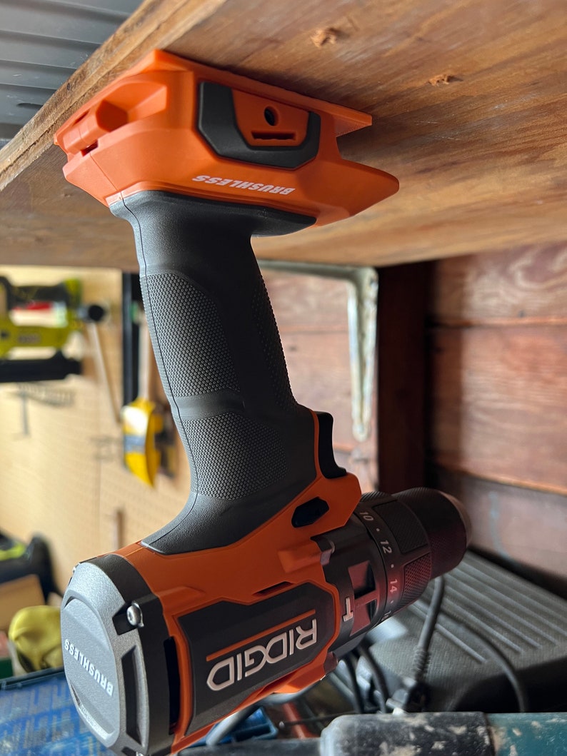 Puede incluir: Un taladro inal&aacute;mbrico naranja y negro con el nombre de la marca "RIDGID" y el texto "BRUSHLESS" en el lateral. El taladro est&aacute; colgado de una estanter&iacute;a de madera.