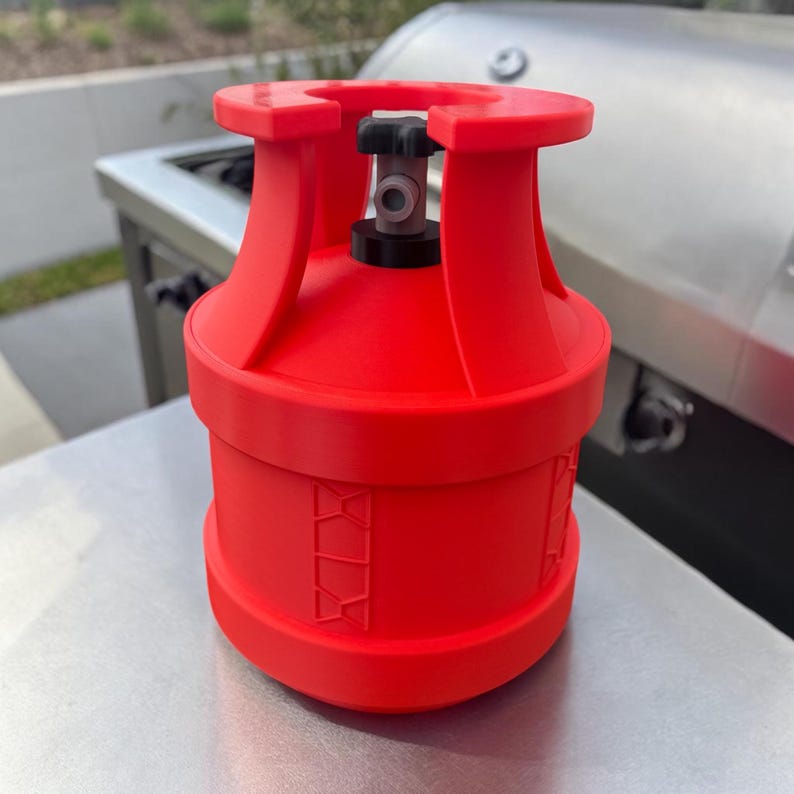May include: A bright red propane tank with a black valve and a silver metal surface in the background. The tank has a cylindrical shape with a rounded top and bottom. The tank has a series of decorative geometric shapes around the middle.