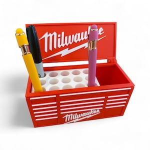 Pu&ograve; includere: Un portapenne rosso a forma di cassetta degli attrezzi con il logo "Milwaukee" in bianco. Contiene una penna gialla, un pennarello nero e una penna rosa. La cassetta degli attrezzi ha strisce bianche e un interno bianco con pi&ugrave; fori per penne.