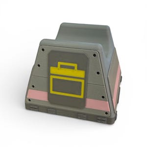 以下が含まれることがあります： 前面に黄と灰色の長方形のデザインが施された、灰色の幾何学的なオブジェクト。オブジェクトにはピンクのストライプがあり、上部にハンドル状の形状があります。デザインはツールボックスまたはコンテナを連想させます。