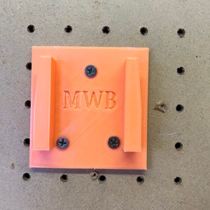 Puede incluir: Soporte de pared de pl&aacute;stico naranja con las letras "MWB" impresas en &eacute;l. Hay tres tornillos que sujetan el soporte a la pared.