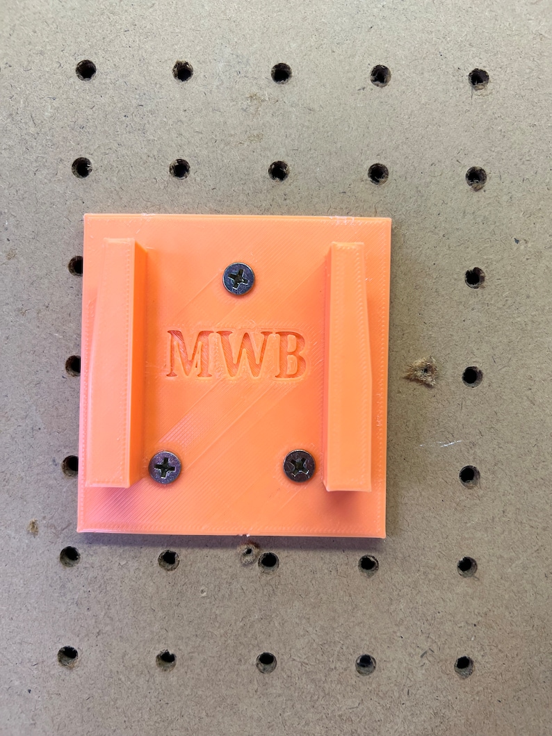 Puede incluir: Soporte de pared de pl&aacute;stico naranja con las letras "MWB" impresas en &eacute;l. Hay tres tornillos que sujetan el soporte a la pared.
