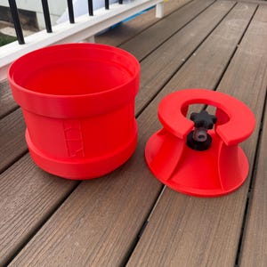 May include: Bright red plastic bucket and lid. The bucket is cylindrical with a wide opening and a slightly flared base. The lid has a central knob and a flared, conical shape. The items are on a wooden deck.