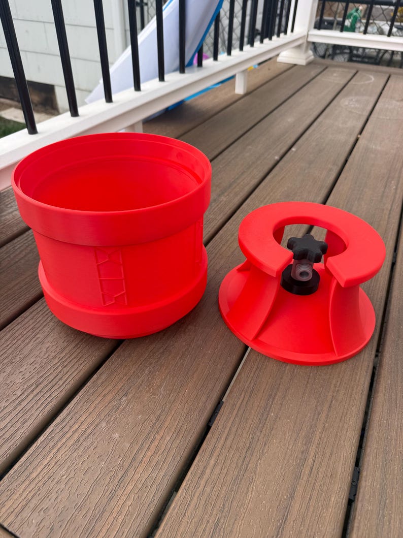 May include: Bright red plastic bucket and lid. The bucket is cylindrical with a wide opening and a slightly flared base. The lid has a central knob and a flared, conical shape. The items are on a wooden deck.
