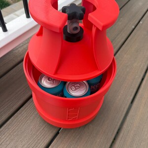 May include: A red, cylindrical cooler with a black valve and star-shaped handle. The cooler holds several blue cans. The cooler has a unique design and is sitting on a wooden deck.