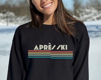 Könnte beinhalten: Ein schwarzer Pullover mit dem Text "APRÈS SKI" in einer Retro-Schriftart mit bunten Streifen darunter. Das Model trägt eine weiße Strickmütze mit Bommel.