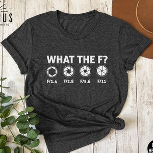 Könnte beinhalten: Dunkelgraues T-Shirt mit einem weißen Grafikdruck von Kamera-Blendenstufen. Der Text "WHAT THE F?" befindet sich über der Grafik. Die Grafik zeigt vier verschiedene Blendenstufen: F/1.4, F/2.8, F/5.6 und F/11.