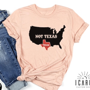 May include: A peach-colored t-shirt with a black and red graphic of the United States with Texas highlighted in red. The text "Not Texas" is printed above Texas. The shirt is laid flat on a white background with a pair of blue jeans.