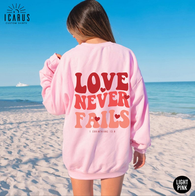 Puede incluir: Una sudadera rosa claro con el texto "LOVE NEVER FAILS" en una fuente retro con corazones. El texto est&aacute; en tonos de rojo y rosa. La sudadera la lleva puesta una persona de pie en una playa.