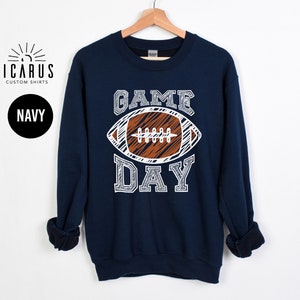 以下が含まれることがあります： 白と茶色のフットボールのグラフィックと「GAME DAY」の文字が入ったネイビーブルーのスウェットシャツ。スウェットシャツはクルーネックと長袖です。「ICARUS CUSTOM SHIRTS」のブランドロゴが左上にあります。
