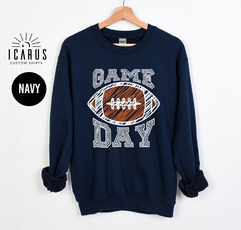 以下が含まれることがあります： 白と茶色のフットボールのグラフィックと「GAME DAY」の文字が入ったネイビーブルーのスウェットシャツ。スウェットシャツはクルーネックと長袖です。「ICARUS CUSTOM SHIRTS」のブランドロゴが左上にあります。