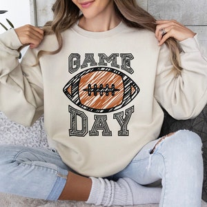 以下が含まれることがあります： クリーム色のスウェットシャツ。中央に黒いアウトラインのフォントで「GAME DAY」の文字とフットボールのグラフィックが描かれています。フットボールは茶色で、白い紐と黒いアウトラインがあります。カジュアルで快適なスタイルです。
