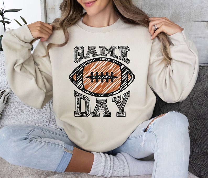 以下が含まれることがあります： クリーム色のスウェットシャツ。中央に黒いアウトラインのフォントで「GAME DAY」の文字とフットボールのグラフィックが描かれています。フットボールは茶色で、白い紐と黒いアウトラインがあります。カジュアルで快適なスタイルです。