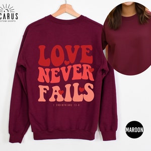Puede incluir: Sudadera color granate con el texto "LOVE NEVER FAILS" en una fuente retro con corazones. El texto "1 Corintios 13:8" est&aacute; impreso debajo del texto principal.